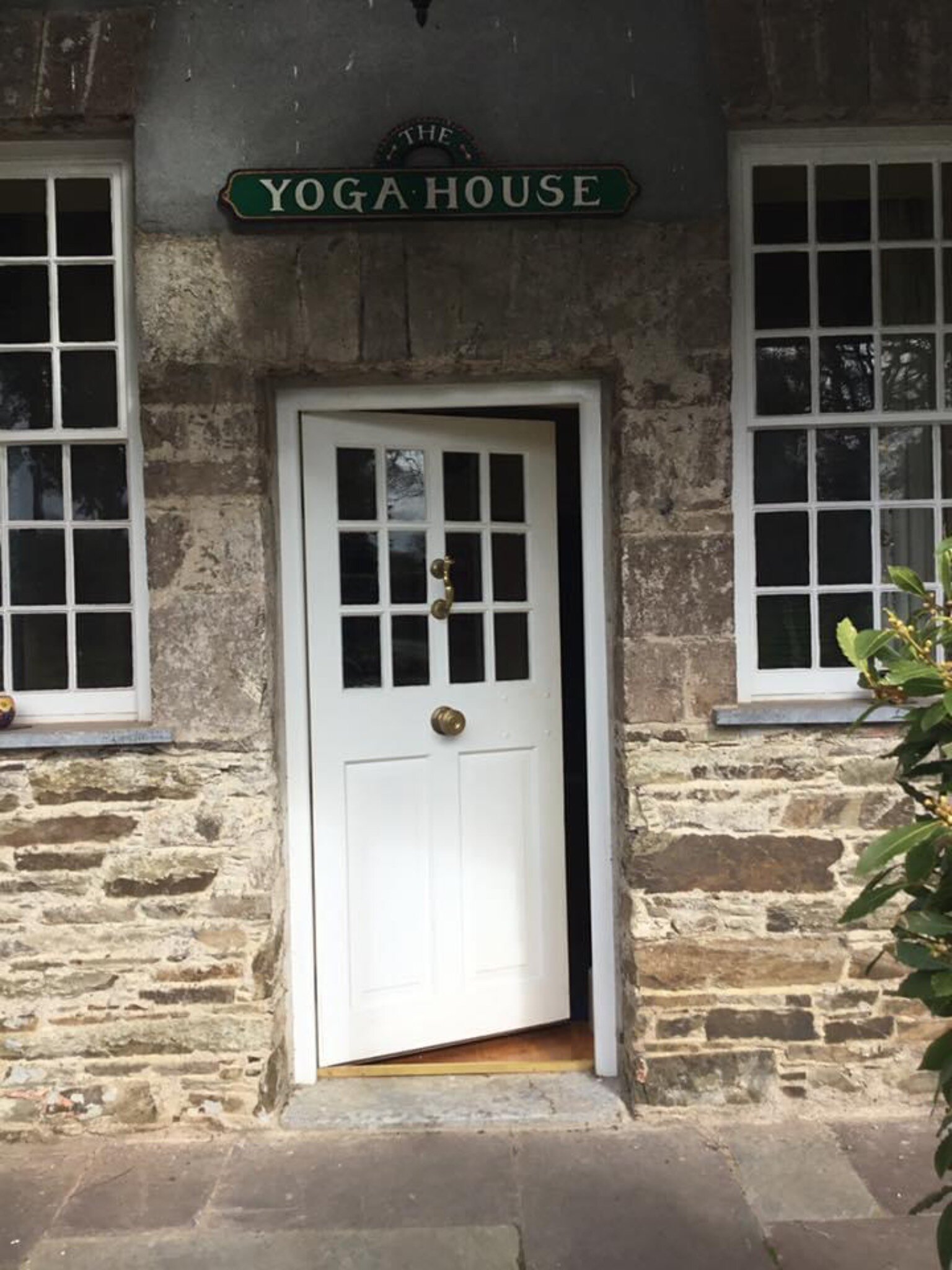 A white front door at an entrance to a stone building. A wooden sign above the door reads, in white handpainted letters, "YOGA HOUSE."